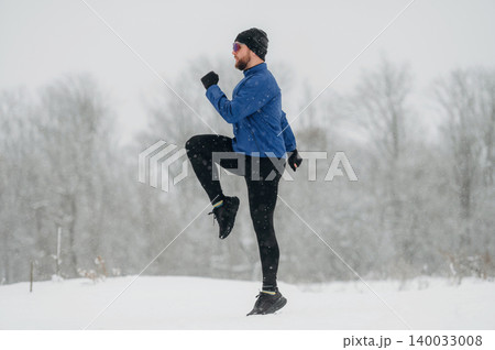 Male runner in blue jacket and black leggings exercises in snowy landscape, snowflakes falling, trees blurred in the background, winter fitness activity 140033008
