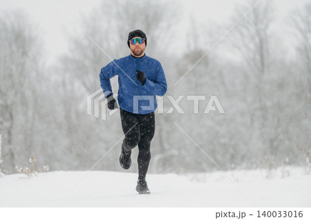 Male runner in blue jacket and black leggings sprinting through snowy landscape during winter, snowflakes falling and trees blurred in the background Male runner in blue jacket and black leggings sprinting through snowy landscape during winter, snowflakes falling and trees blurred in the background 140033016