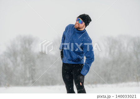 Male athlete wearing blue jacket and sunglasses stretches outdoors in snowy landscape with trees and falling snowflakes in the background 140033021