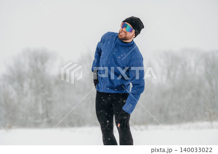 Male athlete wearing blue jacket and sunglasses stretches during winter workout in snowy landscape with trees and soft snowfall in the background 140033022