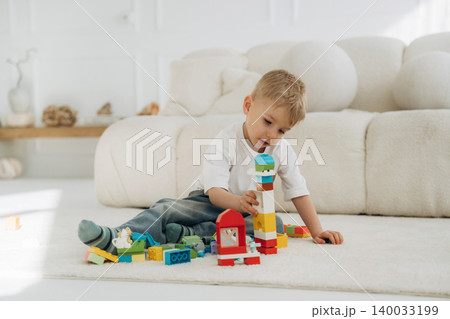 Young boy playing with colorful building blocks on a soft rug in a bright, modern living room with a cozy sofa and decorative items in the background 140033199