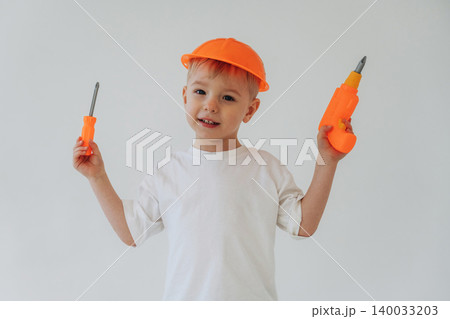 Young boy wearing an orange hard hat holds a toy screwdriver and toy drill, smiling while standing against a plain white background Young boy wearing an orange hard hat holds a toy screwdriver and toy drill, smiling while standing against a plain white background 140033203