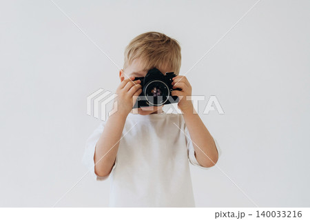 Young boy holding a digital camera in front of his face, wearing a white t-shirt, with a plain light background creating a clean and simple composition 140033216