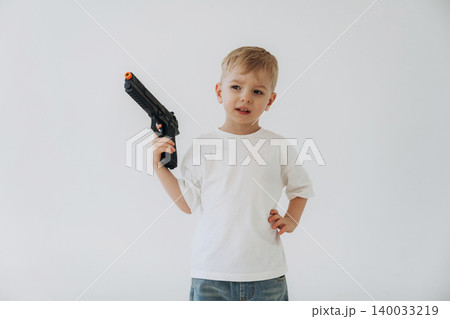 Young boy holding a toy gun in a white t-shirt and denim jeans, standing confidently against a plain white background 140033219