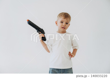 Young boy in white t-shirt and blue jeans holds a toy gun with an orange tip, posing against a plain white background in a playful manner Young boy in white t-shirt and blue jeans holds a toy gun with an orange tip, posing against a plain white background in a playful manner 140033220