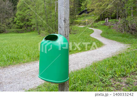 A modern green plastic litter bin mounted on a wooden post next to a scenic gravel walking path in a summer forest park. Concept of environmental care, waste management, and public cleanliness. Waste A modern green plastic litter bin mounted on a wooden post next to a scenic gravel walking path in a summer forest park. Concept of environmental care, waste management, and public cleanliness. Waste 140033242