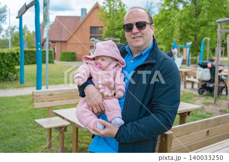 A happy loving father wearing sunglasses holds his small child dressed in a pink warm jumpsuit on an outdoor public playground. Concept of happy parenthood, dad life, and family weekend A happy loving father wearing sunglasses holds his small child dressed in a pink warm jumpsuit on an outdoor public playground. Concept of happy parenthood, dad life, and family weekend 140033250