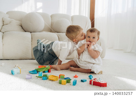 Young boy kisses baby sister while playing with colorful building blocks on soft carpet in a bright, modern living room with large windows Young boy kisses baby sister while playing with colorful building blocks on soft carpet in a bright, modern living room with large windows 140033257