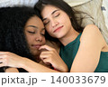 Two young women resting together while holding hands in tender moment of friendship and connection while lying on bed with eyes closed 140033679