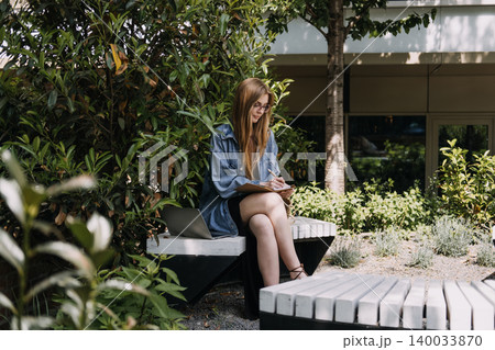 Woman sits on bench in green courtyard writing in notebook next to laptop computer. Green-space remote work, biophilic workplace design, urban integrated office, ecological campus planning. 140033870