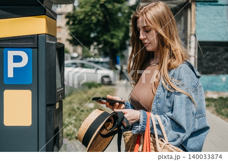 Woman stands next to automated parking terminal holding smartphone near display screen. Cashless city lifestyle, contactless mobile transaction, digital wallet payment, app based billing 140033874