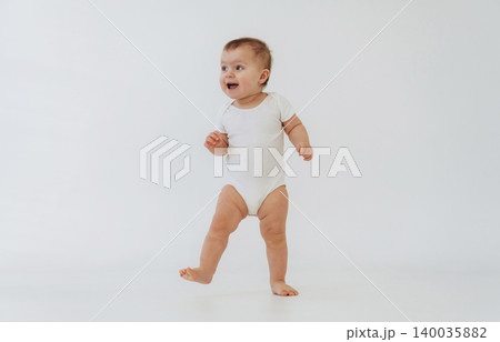Baby wearing white onesie stands on one leg while smiling against a plain white background, showcasing early walking skills and playful demeanor Baby wearing white onesie stands on one leg while smiling against a plain white background, showcasing early walking skills and playful demeanor 140035882