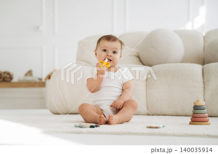 Baby sitting on soft rug in bright room, holding yellow toy, with colorful stacking rings and plush sofa in the background, natural light illuminating the scene Baby sitting on soft rug in bright room, holding yellow toy, with colorful stacking rings and plush sofa in the background, natural light illuminating the scene 140035914