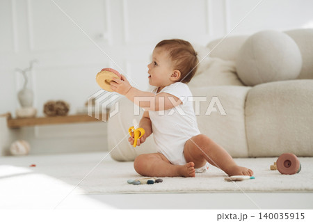 Infant child sitting on a soft rug in a bright room, holding a toy while surrounded by colorful play items and a plush sofa in the background Infant child sitting on a soft rug in a bright room, holding a toy while surrounded by colorful play items and a plush sofa in the background 140035915