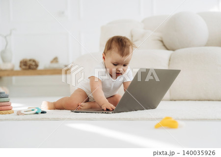Infant child in white onesie interacts with laptop on soft rug in bright living room, with colorful toys scattered nearby and a cozy sofa in the background Infant child in white onesie interacts with laptop on soft rug in bright living room, with colorful toys scattered nearby and a cozy sofa in the background 140035926