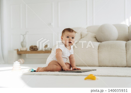 Baby in white onesie sits on soft carpet beside laptop and colorful toys in bright, modern living room with minimalist decor and natural light Baby in white onesie sits on soft carpet beside laptop and colorful toys in bright, modern living room with minimalist decor and natural light 140035931
