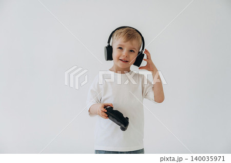 Young boy wearing headphones smiles while holding a video game controller in front of a plain white background, showcasing a playful and engaging gaming moment Young boy wearing headphones smiles while holding a video game controller in front of a plain white background, showcasing a playful and engaging gaming moment 140035971