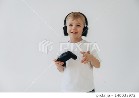 Young boy wearing black headphones holds a video game controller while smiling against a plain white background, showcasing a playful gaming atmosphere Young boy wearing black headphones holds a video game controller while smiling against a plain white background, showcasing a playful gaming atmosphere 140035972