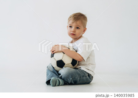 Young boy with short blonde hair wearing a white t-shirt and blue jeans sits on the floor holding a black and white soccer ball against a plain white background 140035985