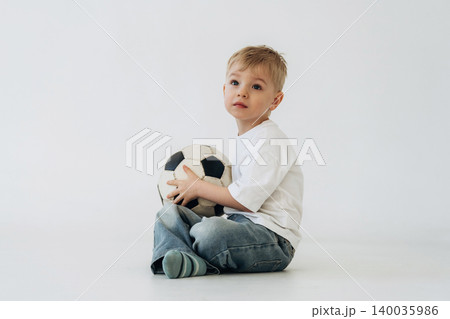 Young boy with short blonde hair wearing a white t-shirt and blue jeans sits on a white floor holding a black and white soccer ball in a bright indoor setting Young boy with short blonde hair wearing a white t-shirt and blue jeans sits on a white floor holding a black and white soccer ball in a bright indoor setting 140035986
