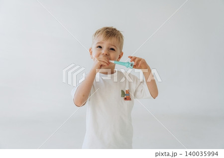 Young boy with light blonde hair wearing a white t-shirt holds a blue toothbrush in front of his mouth, standing against a plain white background Young boy with light blonde hair wearing a white t-shirt holds a blue toothbrush in front of his mouth, standing against a plain white background 140035994