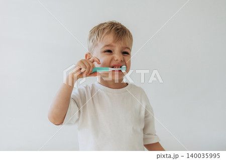 Young boy with light blonde hair brushes his teeth with a blue toothbrush while wearing a white t-shirt against a plain light background Young boy with light blonde hair brushes his teeth with a blue toothbrush while wearing a white t-shirt against a plain light background 140035995