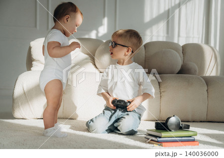 Young Caucasian boy wearing glasses interacts with a baby in a white onesie, surrounded by books and toys on a soft carpet in a bright living room 140036000