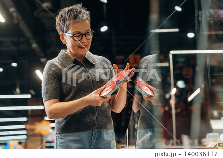 Middle-aged Asian woman with short gray hair wearing glasses examines packaged meat in a grocery store, bright lighting and shelves visible in the background Middle-aged Asian woman with short gray hair wearing glasses examines packaged meat in a grocery store, bright lighting and shelves visible in the background 140036117