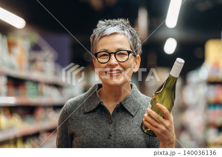Mature woman with short gray hair wearing glasses holds a green wine bottle while smiling in a grocery store aisle filled with various products and shelves Mature woman with short gray hair wearing glasses holds a green wine bottle while smiling in a grocery store aisle filled with various products and shelves 140036136