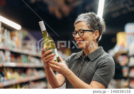 Middle-aged woman with short gray hair wearing glasses examines a bottle of white wine while shopping in a grocery store aisle filled with products Middle-aged woman with short gray hair wearing glasses examines a bottle of white wine while shopping in a grocery store aisle filled with products 140036138