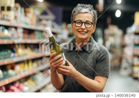 Senior Asian woman with short gray hair holds a bottle of white wine while smiling in a grocery store aisle filled with various food products and shelves Senior Asian woman with short gray hair holds a bottle of white wine while smiling in a grocery store aisle filled with various food products and shelves 140036140