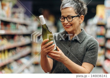 Mature woman with short gray hair wearing glasses examines a bottle of white wine while shopping in a grocery store aisle filled with various products 140036141