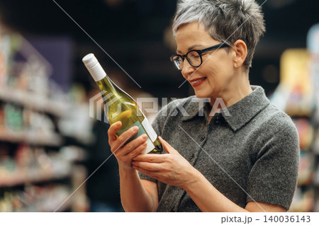 Middle-aged woman with short gray hair wearing glasses examines a bottle of white wine while standing in a well-stocked grocery store aisle filled with various products Middle-aged woman with short gray hair wearing glasses examines a bottle of white wine while standing in a well-stocked grocery store aisle filled with various products 140036143