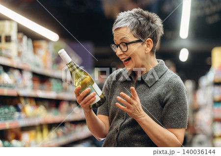 Middle-aged woman with short gray hair and glasses holds a bottle of white wine, smiling in a grocery store aisle filled with various products and shelves 140036144
