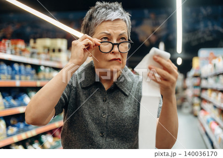 Mature woman with short gray hair wearing glasses examines shopping receipt while standing in grocery store aisle filled with various products and shelves in the background 140036170