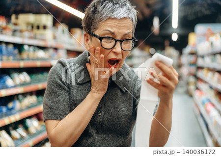 Senior woman with short gray hair and glasses looks shocked while holding a shopping receipt in a grocery store aisle filled with products and shelves 140036172