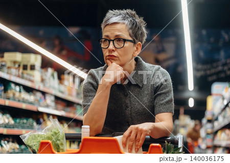 Middle-aged woman with short gray hair and glasses ponders while shopping in a grocery store, with shelves of products and vegetables in the background Middle-aged woman with short gray hair and glasses ponders while shopping in a grocery store, with shelves of products and vegetables in the background 140036175