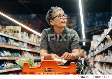 Middle-aged woman with short gray hair and glasses stands by a shopping cart in a grocery store aisle, looking thoughtfully at a shopping list 140036177