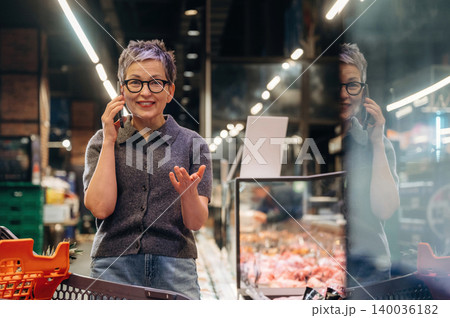 Mature woman with short gray hair talking on a smartphone while gesturing inside a modern grocery store with fresh meat display in the background Mature woman with short gray hair talking on a smartphone while gesturing inside a modern grocery store with fresh meat display in the background 140036182