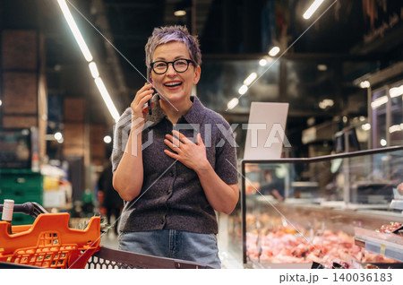 Mature woman with short gray hair talking on a smartphone while standing in a grocery store, smiling and wearing a gray cardigan, with meat display in the background 140036183