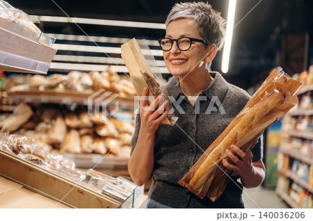 Mature woman with short gray hair wearing glasses holds fresh baguettes in a bakery aisle, surrounded by various types of bread on display in a grocery store 140036206