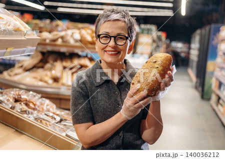 Mature Asian woman with short gray hair wearing glasses holds a loaf of bread in a bakery aisle, surrounded by various baked goods on display Mature Asian woman with short gray hair wearing glasses holds a loaf of bread in a bakery aisle, surrounded by various baked goods on display 140036212