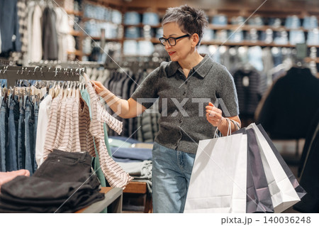 Middle-aged Asian woman shopping for clothes in a retail store, examining a striped shirt while holding shopping bags in a well-organized clothing display Middle-aged Asian woman shopping for clothes in a retail store, examining a striped shirt while holding shopping bags in a well-organized clothing display 140036248