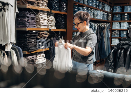 Asian woman in gray sweater examines white clothing item while shopping in a retail store, shelves filled with folded jeans and shirts in the background Asian woman in gray sweater examines white clothing item while shopping in a retail store, shelves filled with folded jeans and shirts in the background 140036256