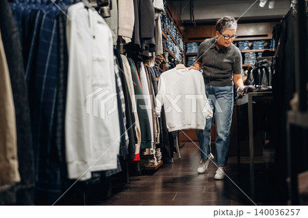 Senior woman shopping for clothing in a retail store, holding a white sweatshirt while browsing through racks of apparel and denim jeans in the background 140036257