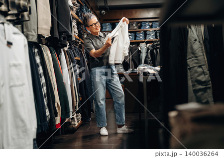 Senior woman with glasses examines white shirt while shopping in clothing store, surrounded by racks of various garments and folded items in the background 140036264