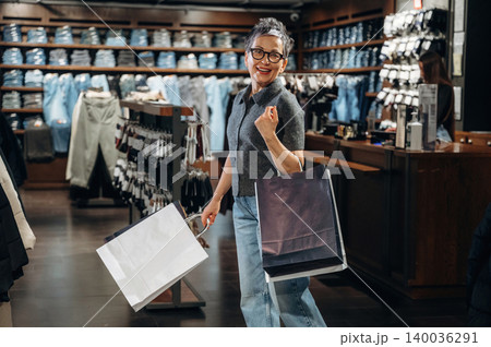 Middle-aged Asian woman in gray sweater and blue jeans smiles while holding shopping bags inside a clothing store with denim apparel displayed in the background Middle-aged Asian woman in gray sweater and blue jeans smiles while holding shopping bags inside a clothing store with denim apparel displayed in the background 140036291