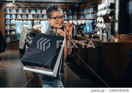 Female shopper with short hair and glasses smiles while holding shopping bags inside a clothing store with denim apparel displayed on shelves in the background 140036309