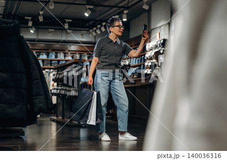 Female shopper with short hair wearing gray shirt and blue jeans holds smartphone while standing in clothing store with shopping bags in hand and racks of clothes in background Female shopper with short hair wearing gray shirt and blue jeans holds smartphone while standing in clothing store with shopping bags in hand and racks of clothes in background 140036316