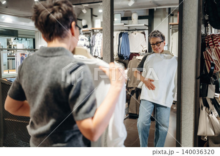 Woman with short hair wearing glasses examines white shirt in front of mirror inside clothing store, casual attire and fashion items displayed in the background 140036320